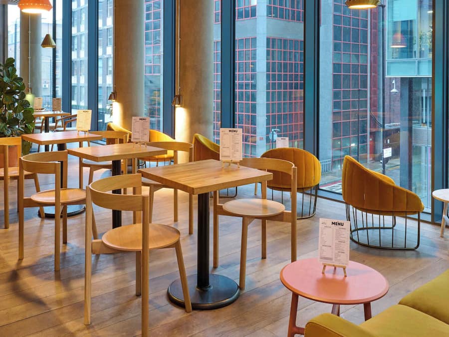 Un café moderno con mesas y sillas de madera, atriles con la carta y grandes ventanales que ofrecen vistas de la ciudad.