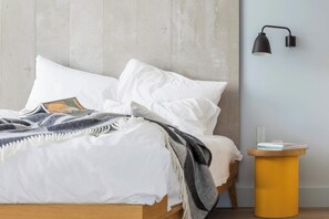 A bed with white bedding, a gray and white throw blanket, and a wooden headboard. A black wall-mounted lamp and a small wooden side table with a book are visible.