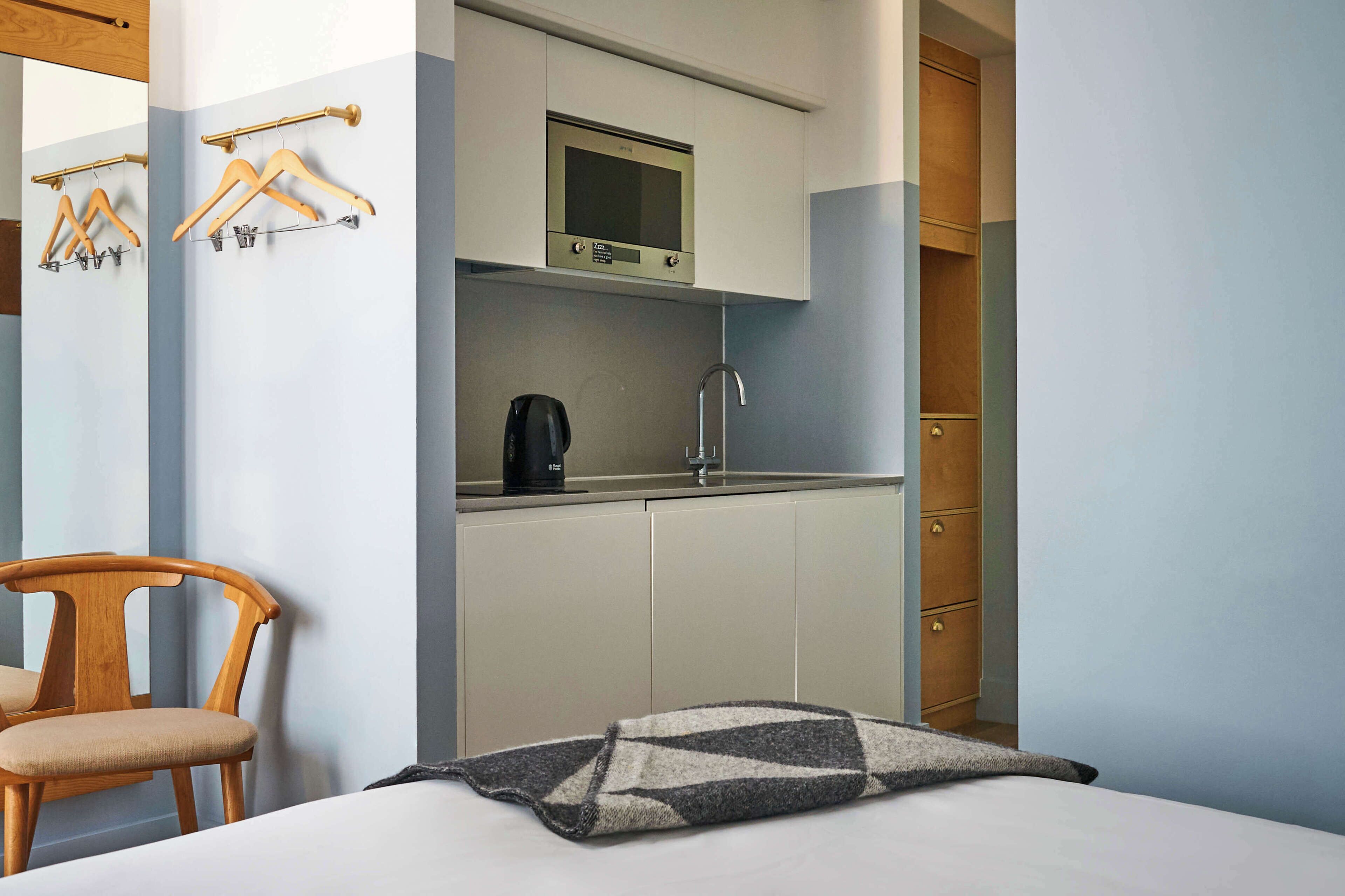 A compact room with a bed, a wooden chair, a kitchenette with a microwave, and a wardrobe.