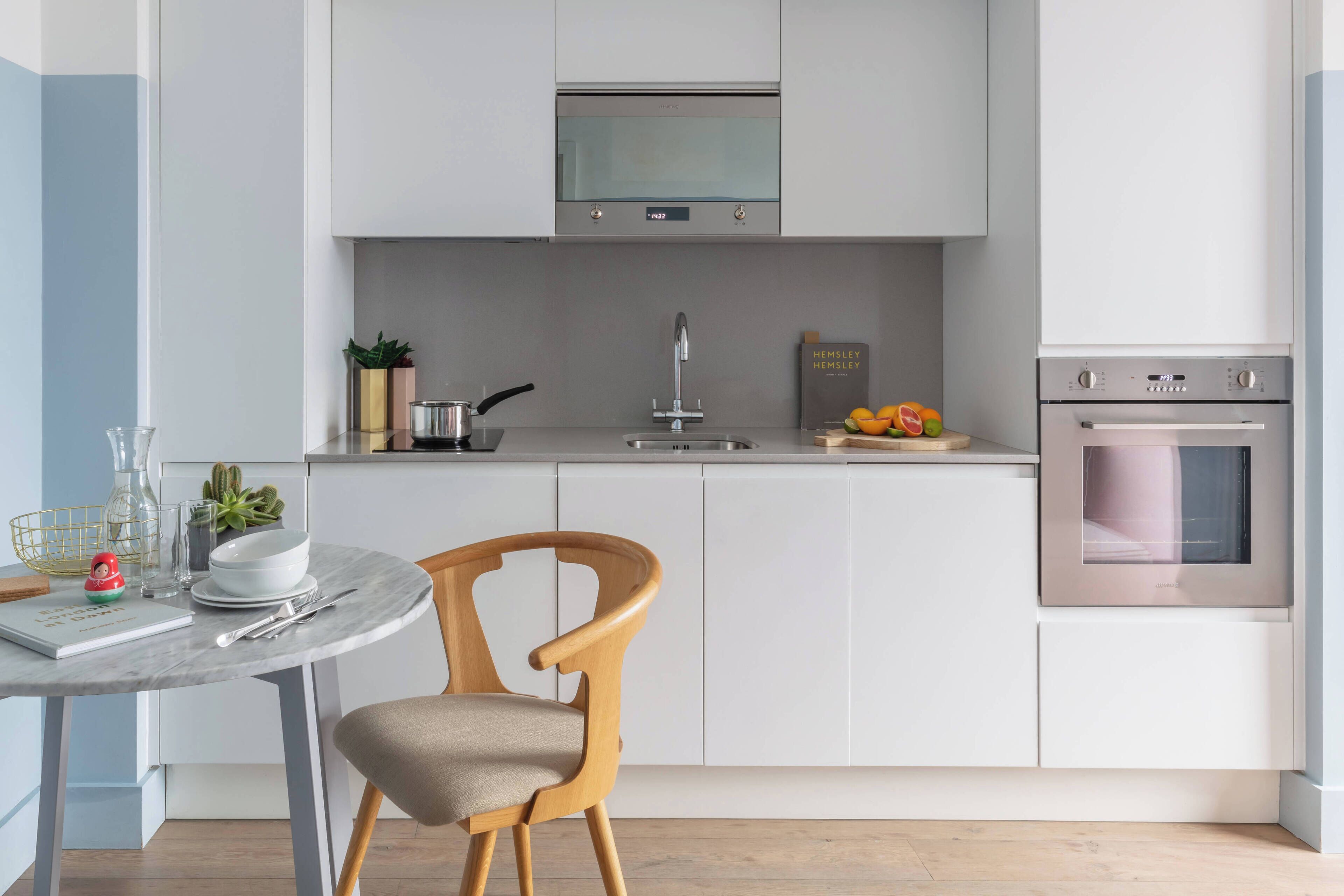 A modern kitchen with white cabinetry, a stainless steel oven, a microwave, and a dining area with a round table and a wooden chair.