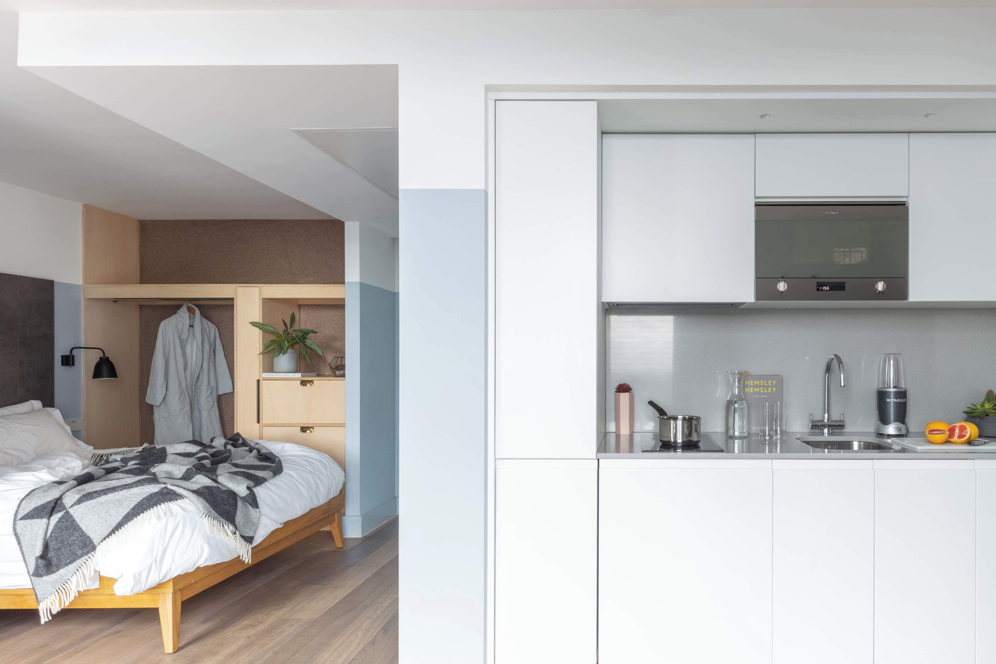 A modern bedroom with a bed, bedside table, and a small plant. Adjacent is a kitchen with a microwave, sink, and countertop.