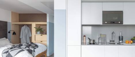 A modern bedroom with a bed, bedside table, and a small plant. Adjacent is a kitchen with a microwave, sink, and countertop.
