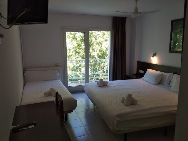 Habitación de hotel con dos camas, ventilador de techo, televisión y balcón con vistas a árboles.