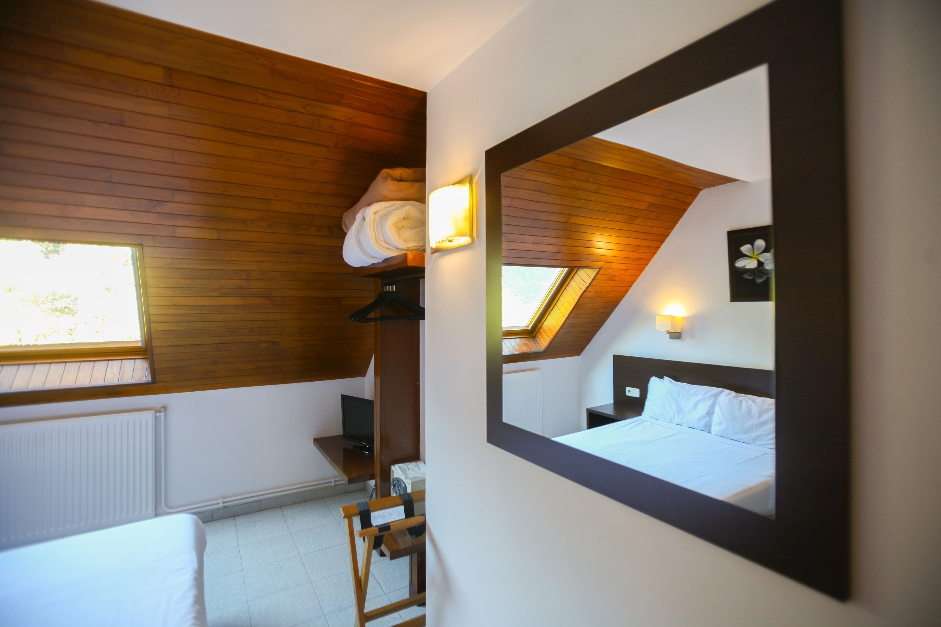 A bedroom with a wooden ceiling, a skylight, a bed, a nightstand, a chair, and a wall-mounted mirror.