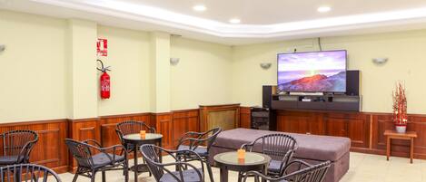Un hall de hotel con sillas de mimbre, un televisor de pantalla plana, un extintor y una pequeña mesa con una vela.
