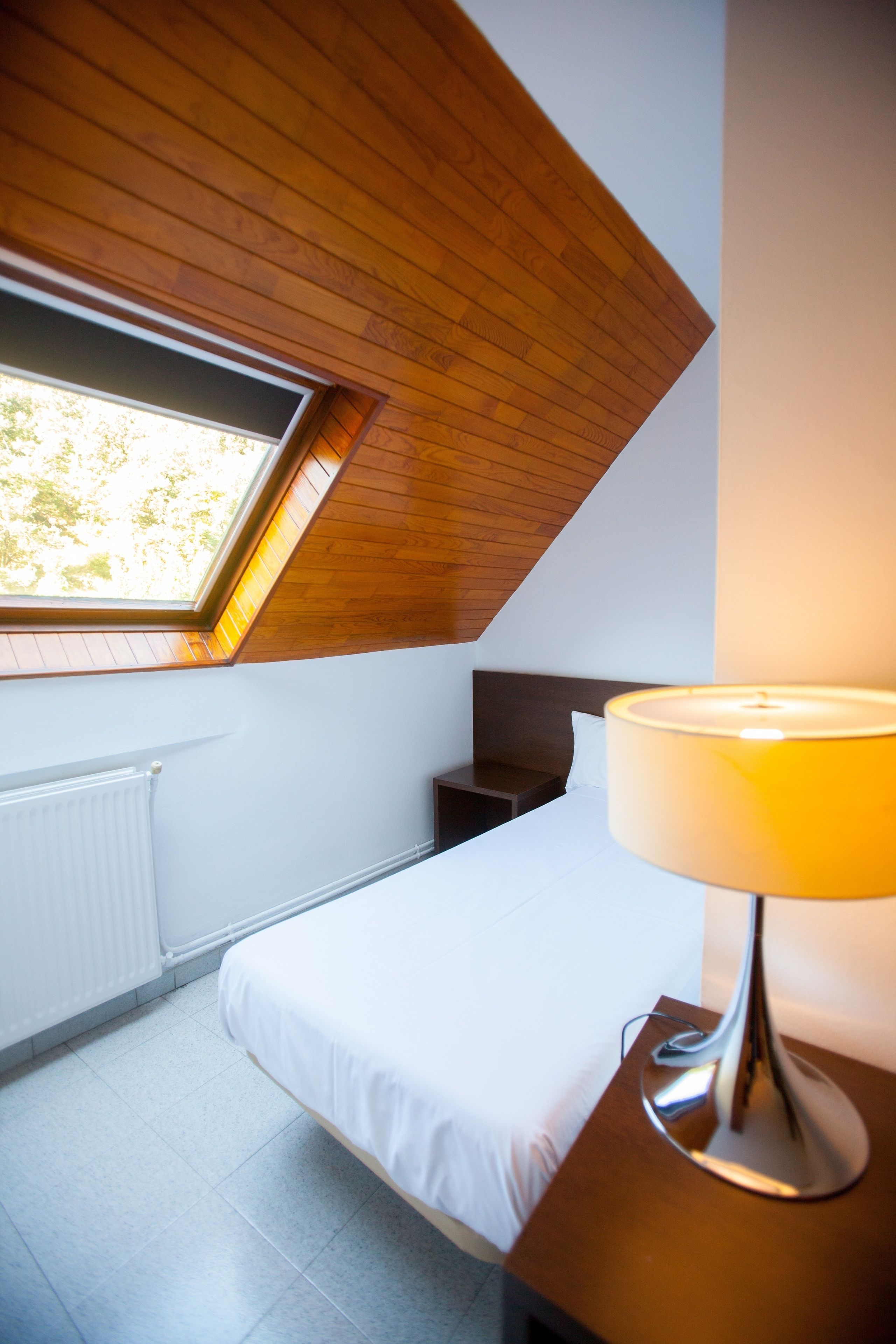 A bedroom with a skylight, a bed, a bedside table with a lamp, and a radiator.
