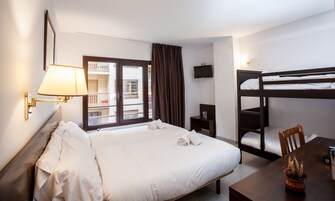 Habitación de hotel con cama, escritorio, literas, televisión y ventana con vista a edificios.