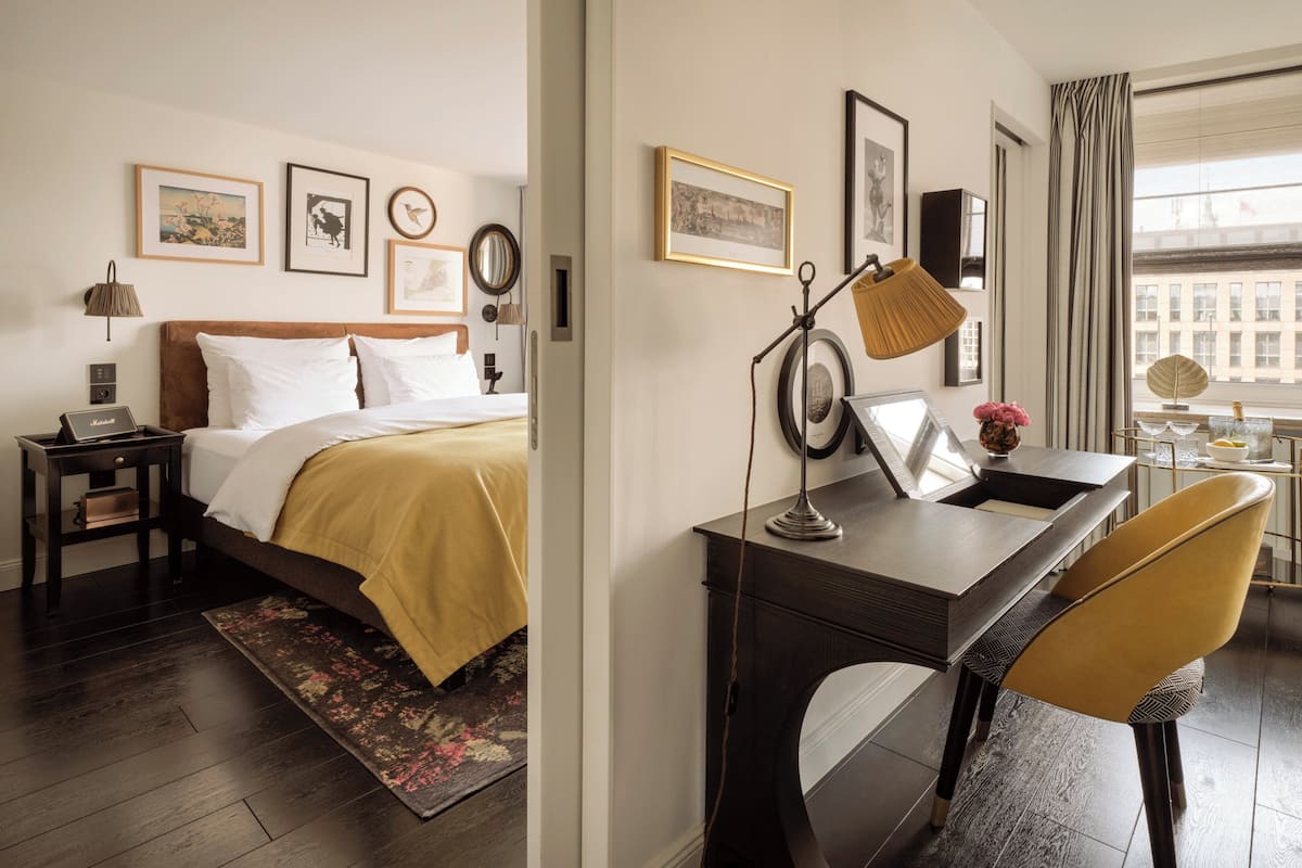 Une chambre d’hôtel comprenant un lit, une table de chevet, une lampe, un bureau, une chaise et des tableaux encadrés accrochés au mur.