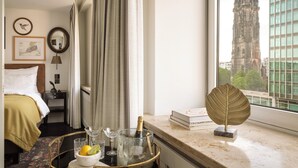 Une chambre d’hôtel avec un lit, une table de chevet, un miroir, une fenêtre donnant sur la ville, une table avec une bouteille de champagne, des verres et une feuille décorative.