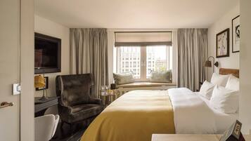 A hotel room with a bed, a chair, a desk with a lamp, a television, and a window with curtains.