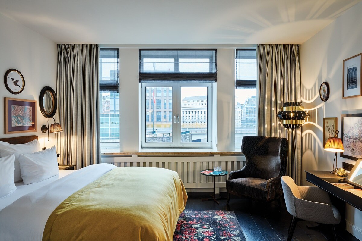 Una camera d'albergo con un letto grande, una scrivania, una sedia, una lampada e vista sulla città dalla finestra.