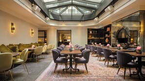 Un hall d’hôtel spacieux, doté d’un plafond haut et d’une verrière, comprenant un espace salon central avec des canapés et des tables, entouré de fauteuils et agrémenté d’un tapis à motifs.