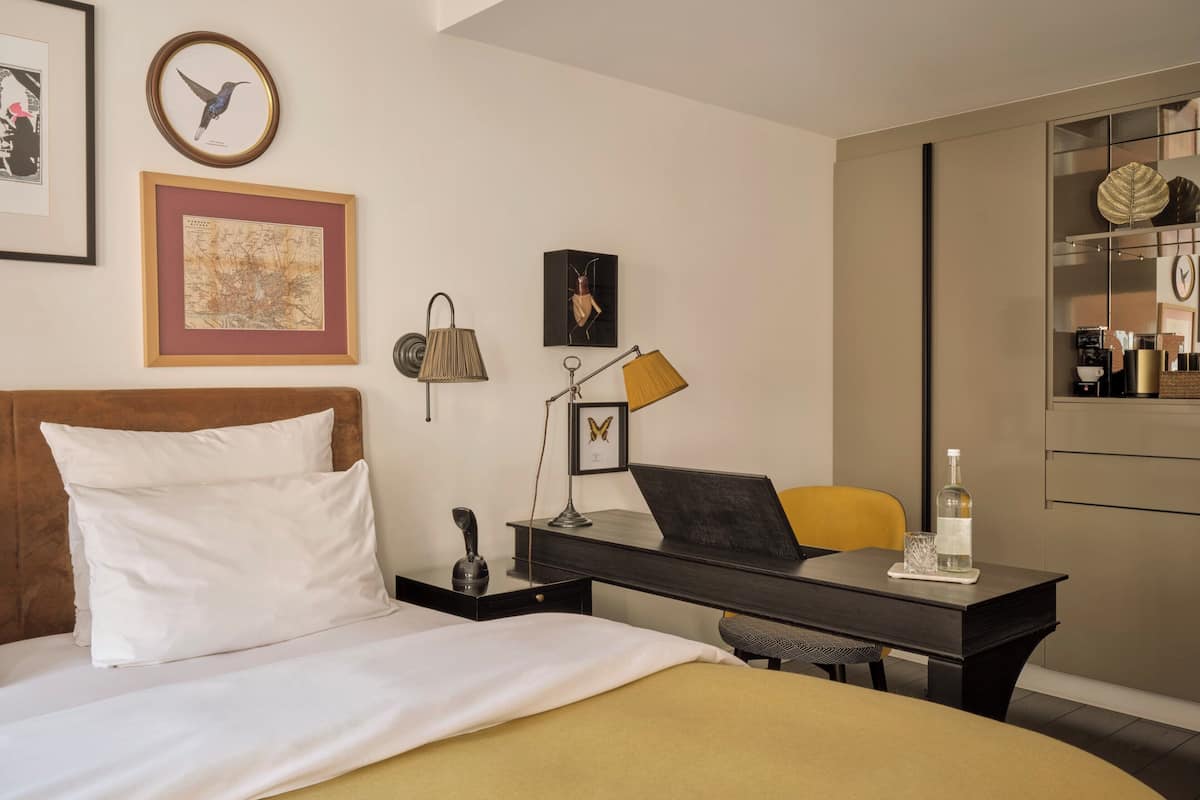 Une chambre d’hôtel avec un lit, un bureau avec un ordinateur portable, une chaise, une lampe et une petite table sur laquelle se trouve une bouteille.