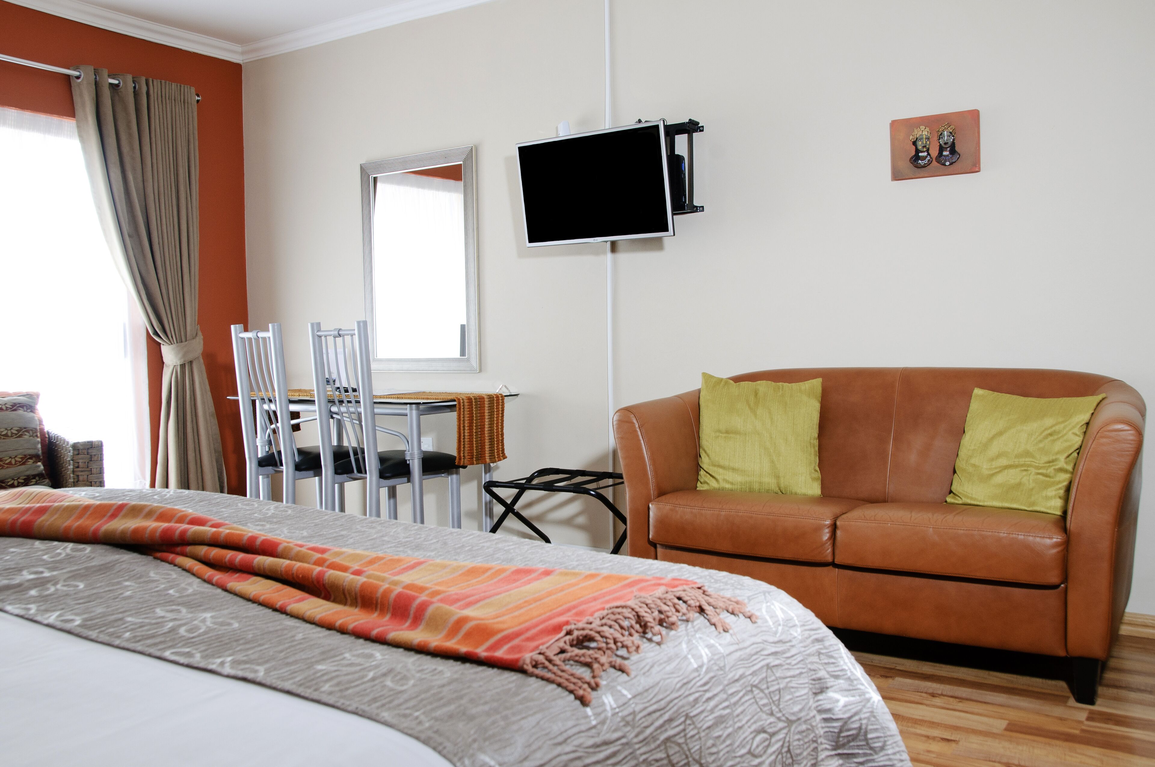 A hotel room with a bed, a brown leather sofa, a television mounted on the wall, and a small table with chairs.