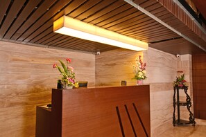 A reception desk with a wooden finish, two flower arrangements, and a modern ceiling with wooden slats and a linear light fixture. - Visitel - A Boutique Hotel (Kolkata)