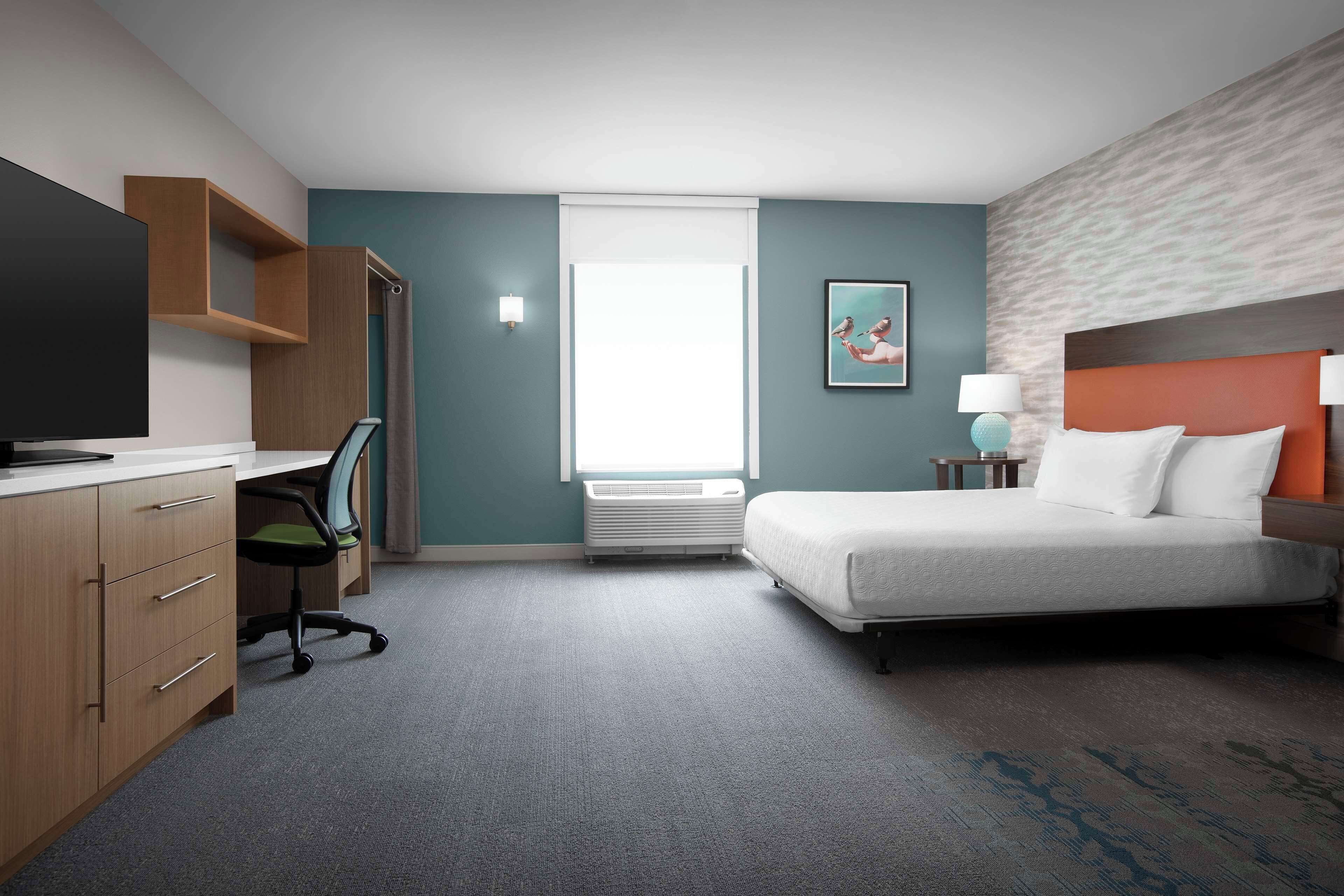 A hotel room with a bed, a desk with a TV, a chair, a window, and a painting on the wall.