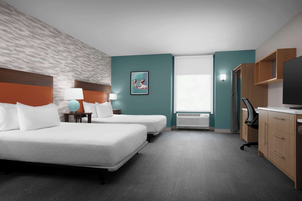 Une chambre d’hôtel avec deux lits, un bureau, une chaise, une télévision, une fenêtre et un radiateur fixé au mur.