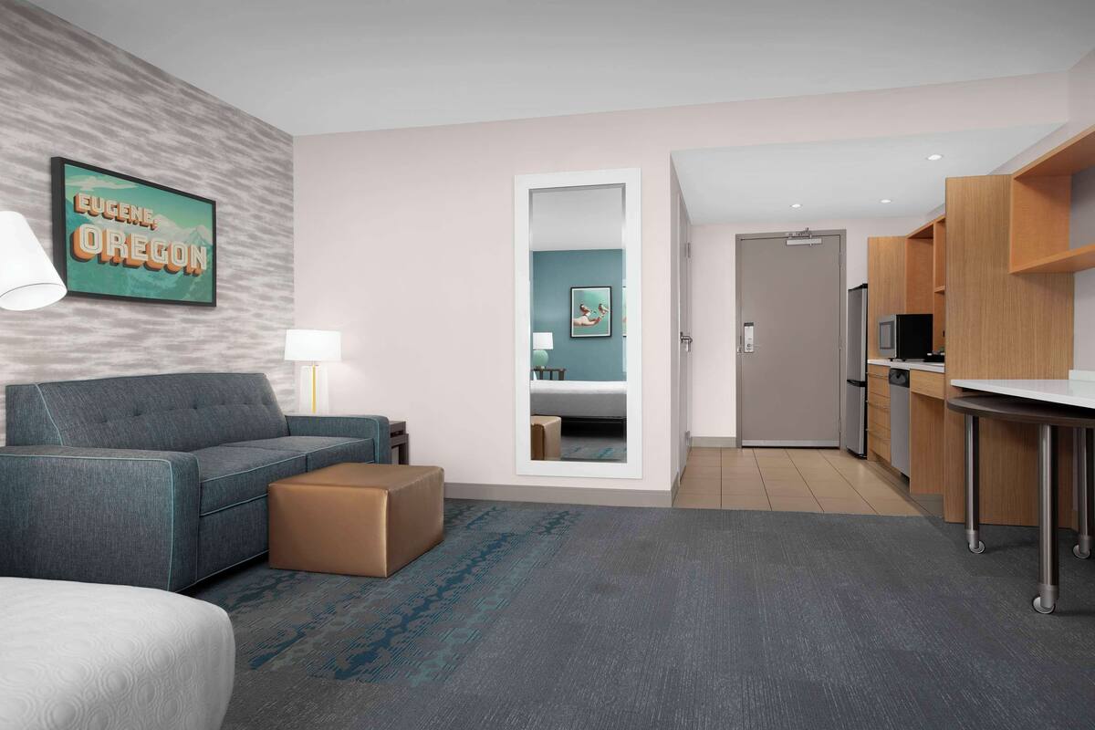 Une chambre d’hôtel moderne avec un canapé gris, un pouf marron, un tableau encadré dans un cadre turquoise, et un coin cuisine doté de meubles en bois et d’un réfrigérateur en acier inoxydable.