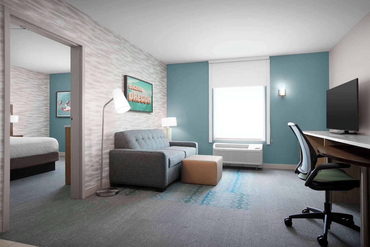 Une chambre d’hôtel avec un lit, un canapé gris, un petit pouf, un bureau avec une chaise, une télévision et une fenêtre avec des stores.