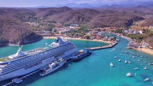 Un grand navire de croisière a accosté dans un port doté d’une marina où étaient amarrés de plus petits bateaux, le tout étant entouré d’une ville côtière et d’un paysage vallonné.