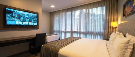 Habitación de hotel con una cama grande, un escritorio, una silla, un televisor montado en la pared y un ventanal con cortinas.