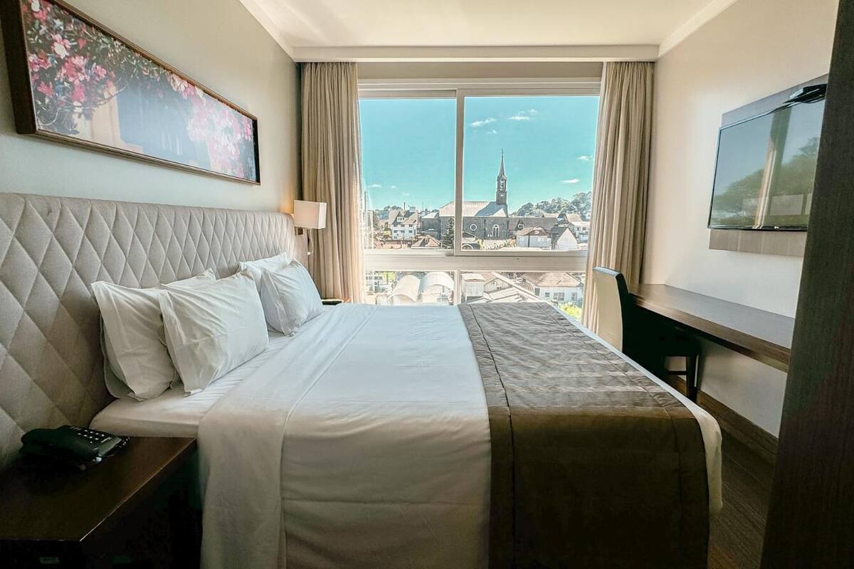 Habitación de hotel con cama, televisión, ventana con vista a la ciudad y un cuadro en la pared.