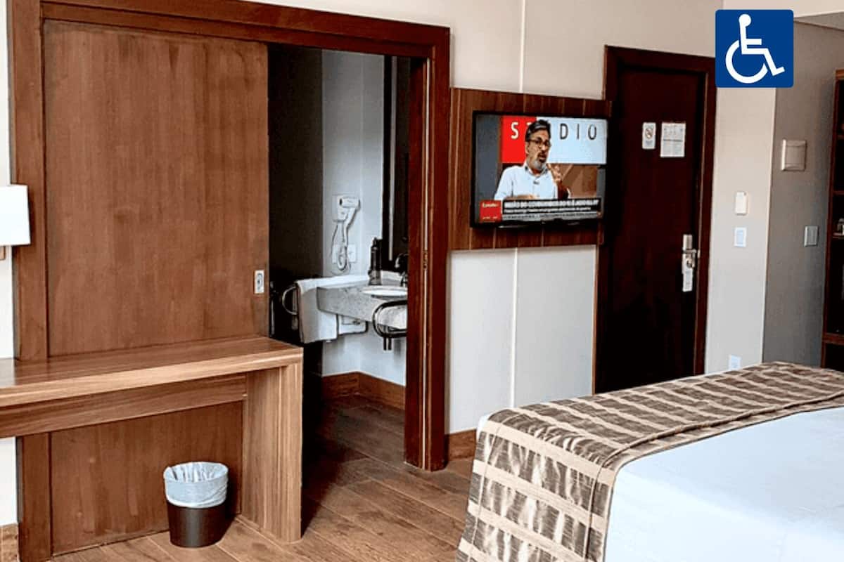 Habitación de hotel con escritorio de madera, televisor montado en la pared, baño con lavamanos y espejo, y señal de accesibilidad para sillas de ruedas.