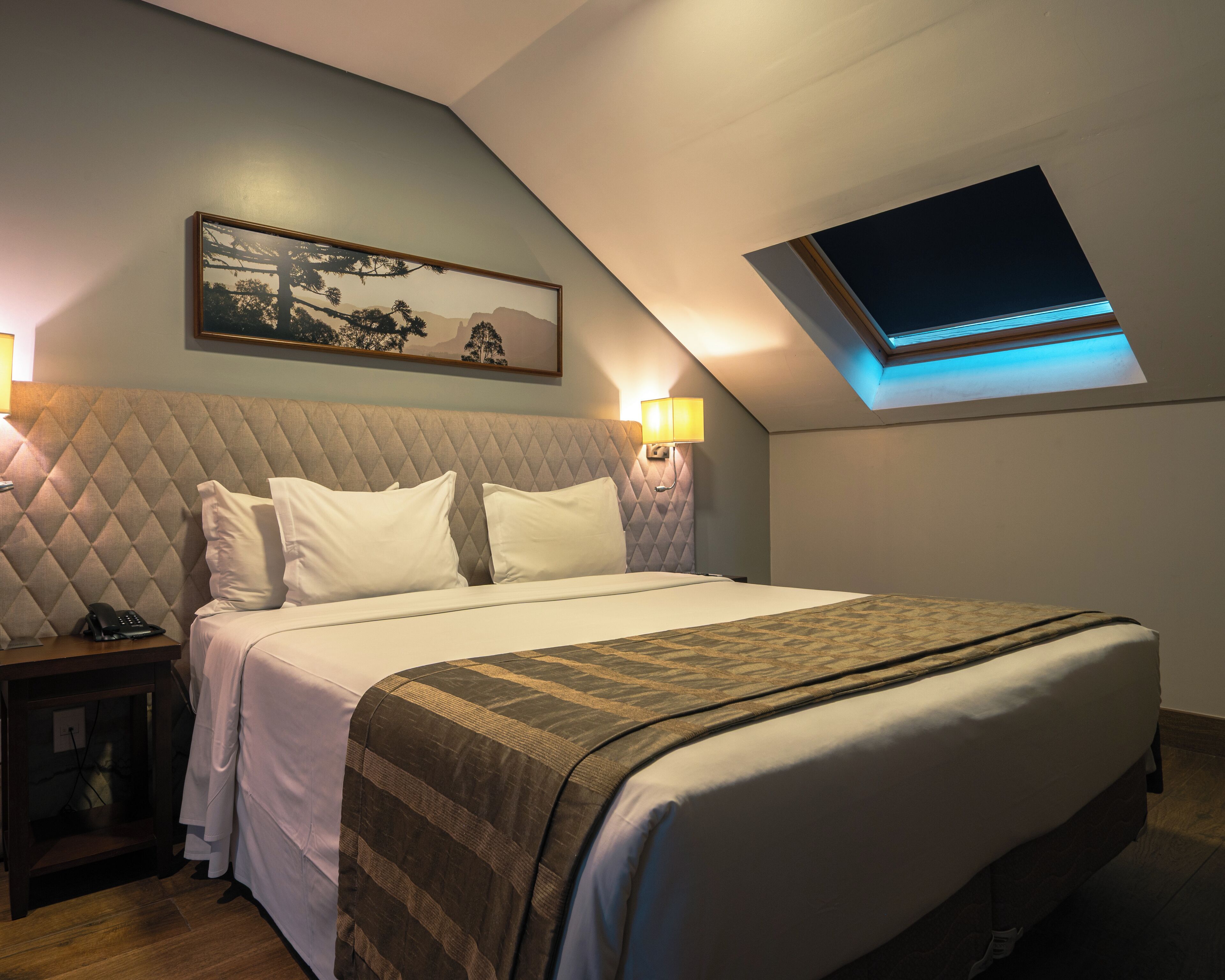 Une chambre d’hôtel avec un lit, une table de chevet, un tableau et une lucarne.