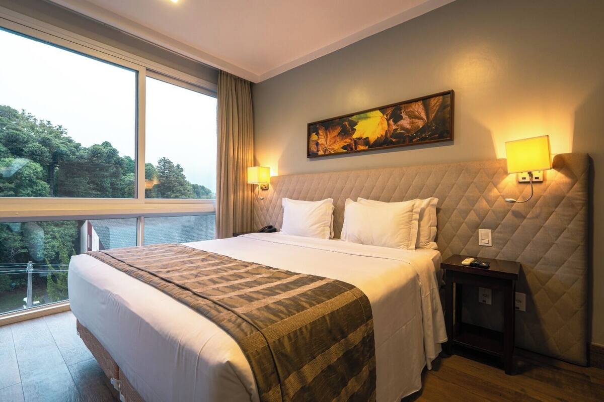 Un cuarto de hotel con una cama grande, vista a árboles por la ventana y un cuadro en la pared.
