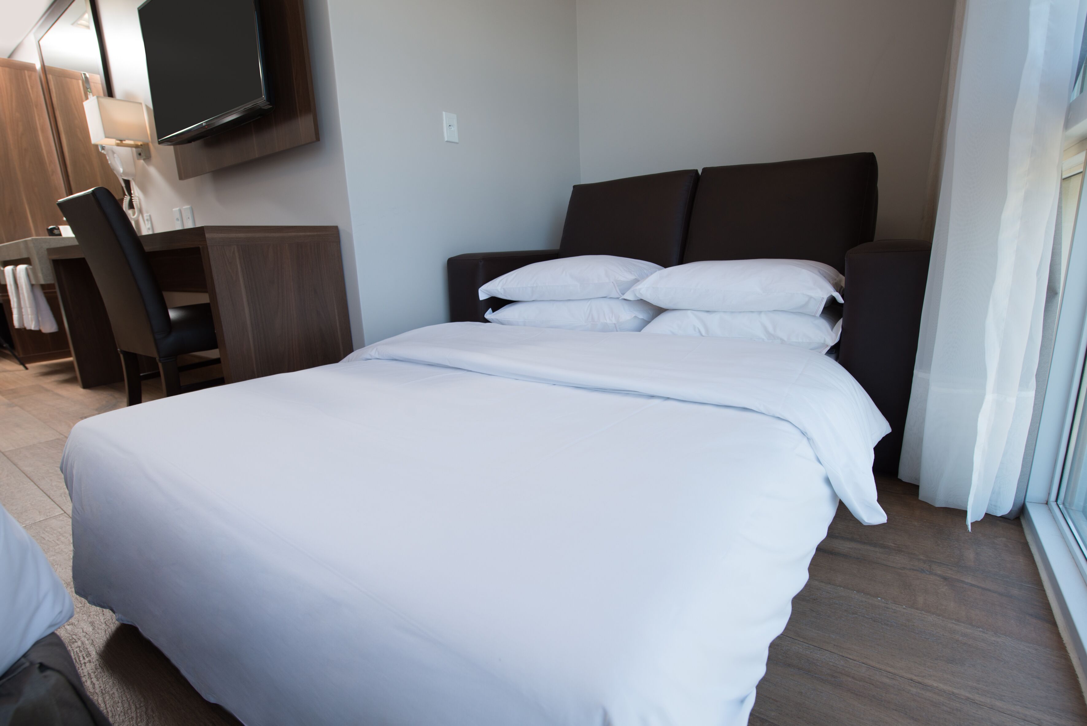 A hotel room with a large bed, a desk with a chair, a flat-screen TV mounted on the wall, and a window with curtains.