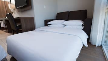 A hotel room with a large bed, a desk with a chair, a flat-screen TV mounted on the wall, and a window with curtains.