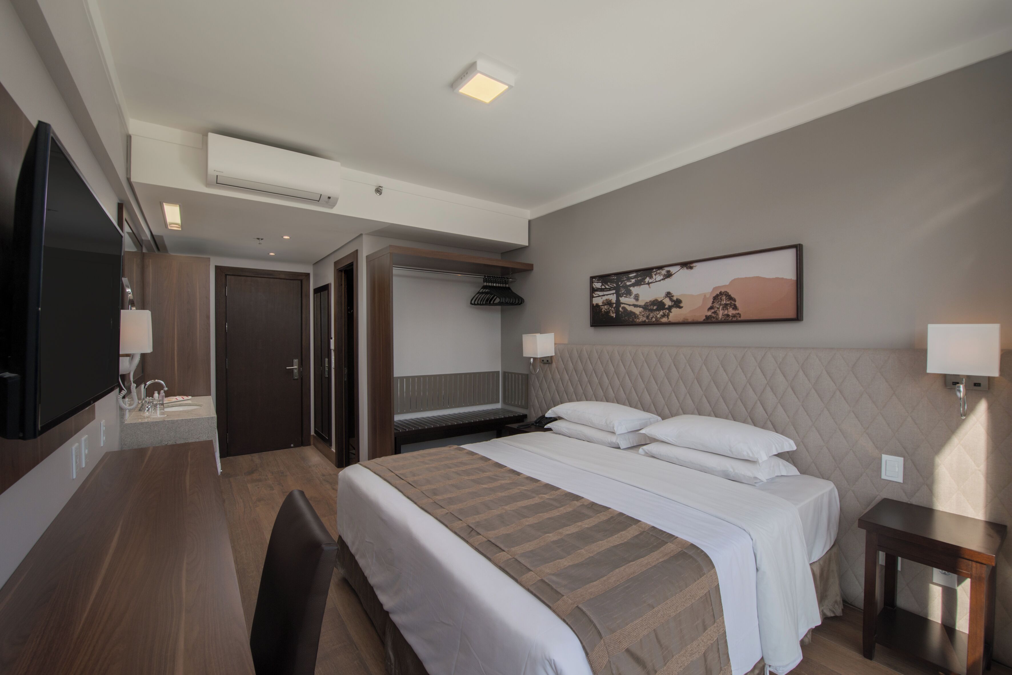 A hotel room with a large bed, a desk with a chair, a flat-screen TV, a bedside table, and a painting on the wall.