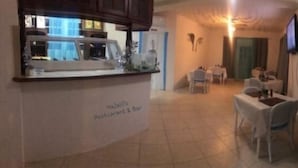 Un espace comprenant un restaurant et un bar, avec une cuisine visible sur le côté gauche, plusieurs tables et chaises disposées pour les repas, et un téléviseur fixé au mur.
