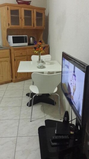Ein kleiner Essbereich mit einem weißen Tisch, Stühlen, einer Blumenvase, einer Mikrowelle und einem Fernseher auf einem Standfuß.