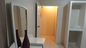 Une chambre d’hôtel avec une coiffeuse blanche, un miroir, un vase en bois, une porte donnant sur un couloir et une armoire contenant des serviettes pliées.