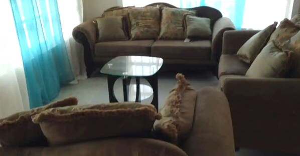 A living room with a brown sofa, a glass coffee table, and a blue curtain. - Mangrove Cay Sea View Villas (Mangrove Cay)