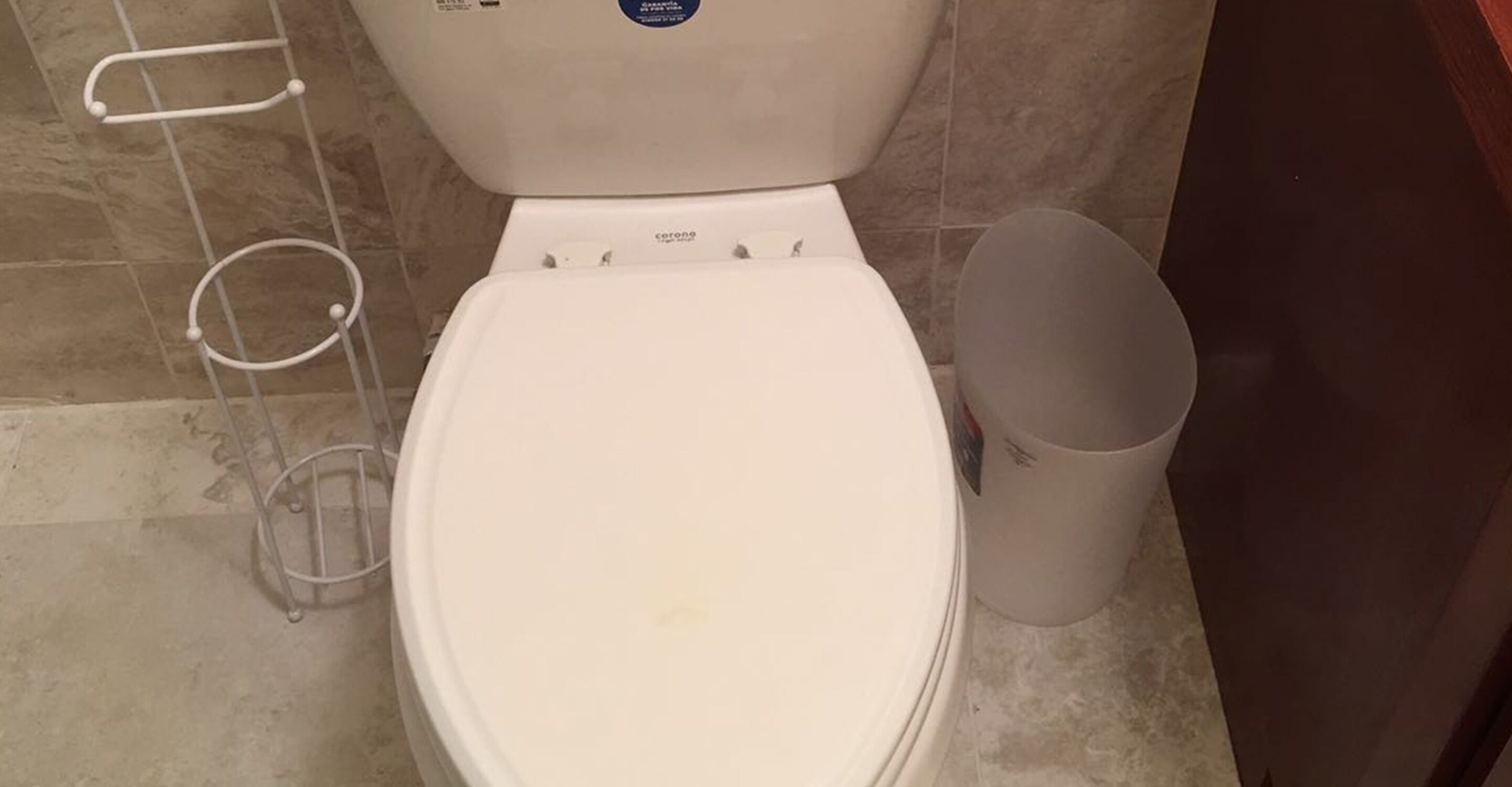 A white toilet with a lid, a toilet paper holder, and a trash can in a bathroom with tiled flooring.