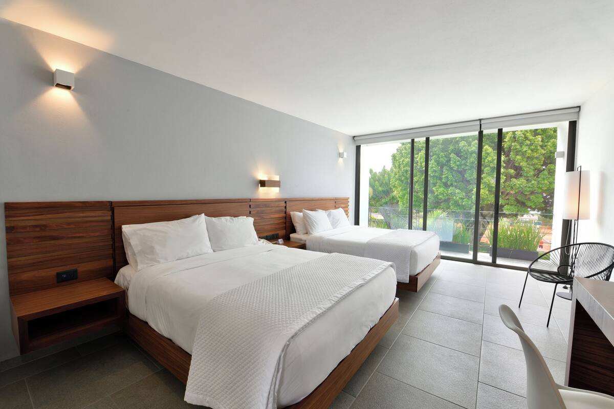 A modern hotel room with two beds, a wooden headboard, a large window with a view of greenery, and a desk with a chair.