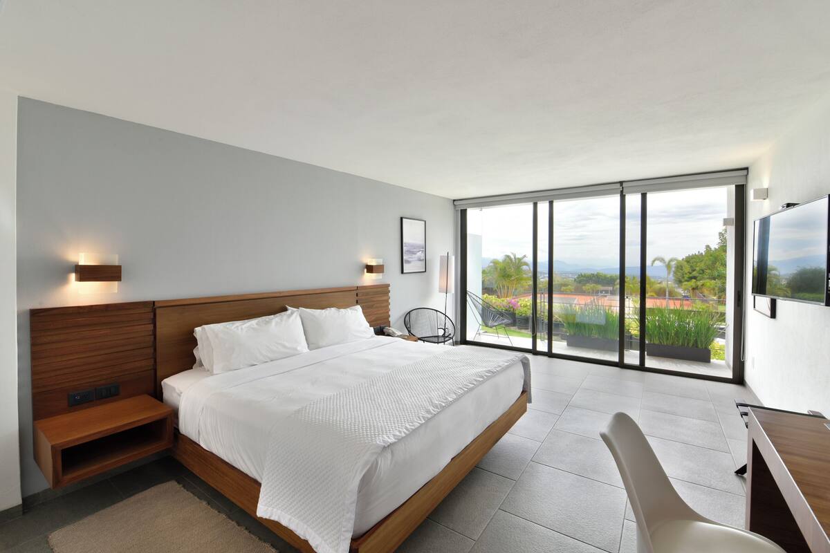 A modern bedroom with a large bed, wooden headboard, bedside tables, a desk, and a chair. It features a large window with a view of greenery and a cityscape.