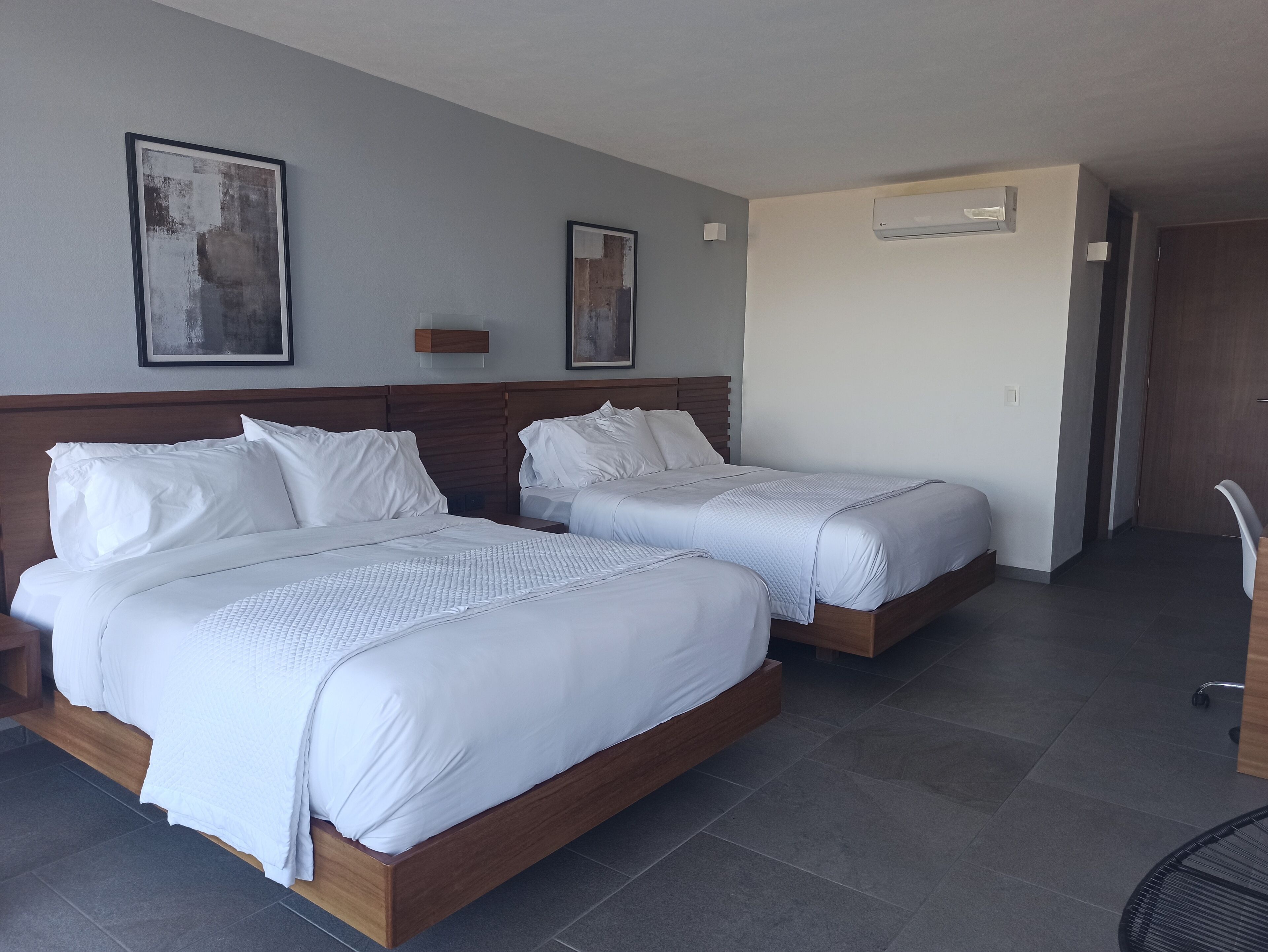 A hotel room with two beds, a wooden headboard, a grey tiled floor, and two abstract paintings on the wall.