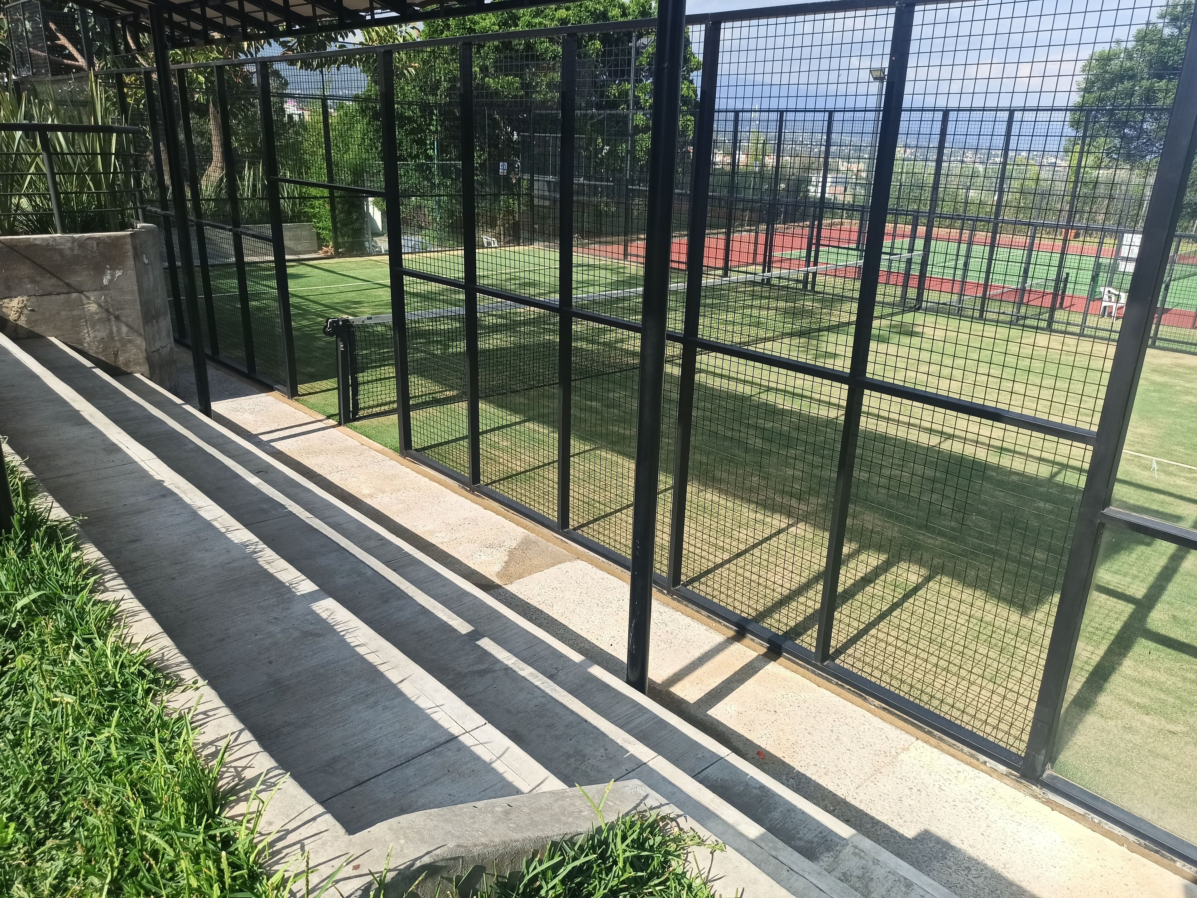 Ein überdachter Außenbereich mit Metallzaun, Betonboden und Blick auf ein Sportfeld.
