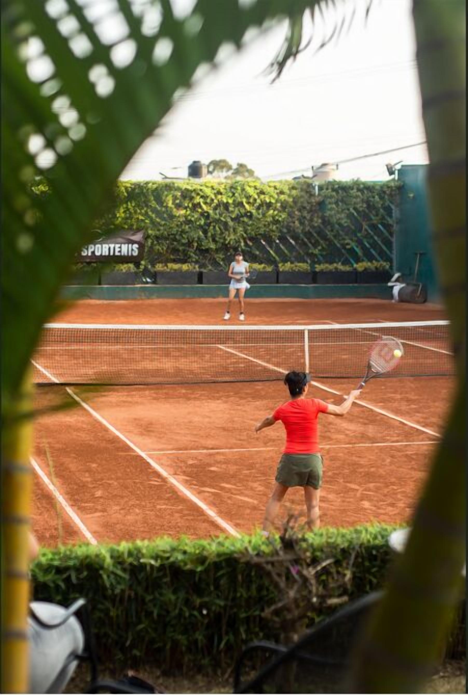 A tennis court with a net in the center, surrounded by greenery and a sign that reads "SPORTENIS".