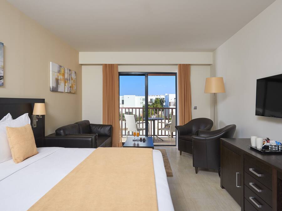 Ein Hotelzimmer mit einem Bett, zwei Sesseln, einem Schreibtisch mit Lampe, einem Fernseher und einem Balkon mit Blick auf Gebäude.