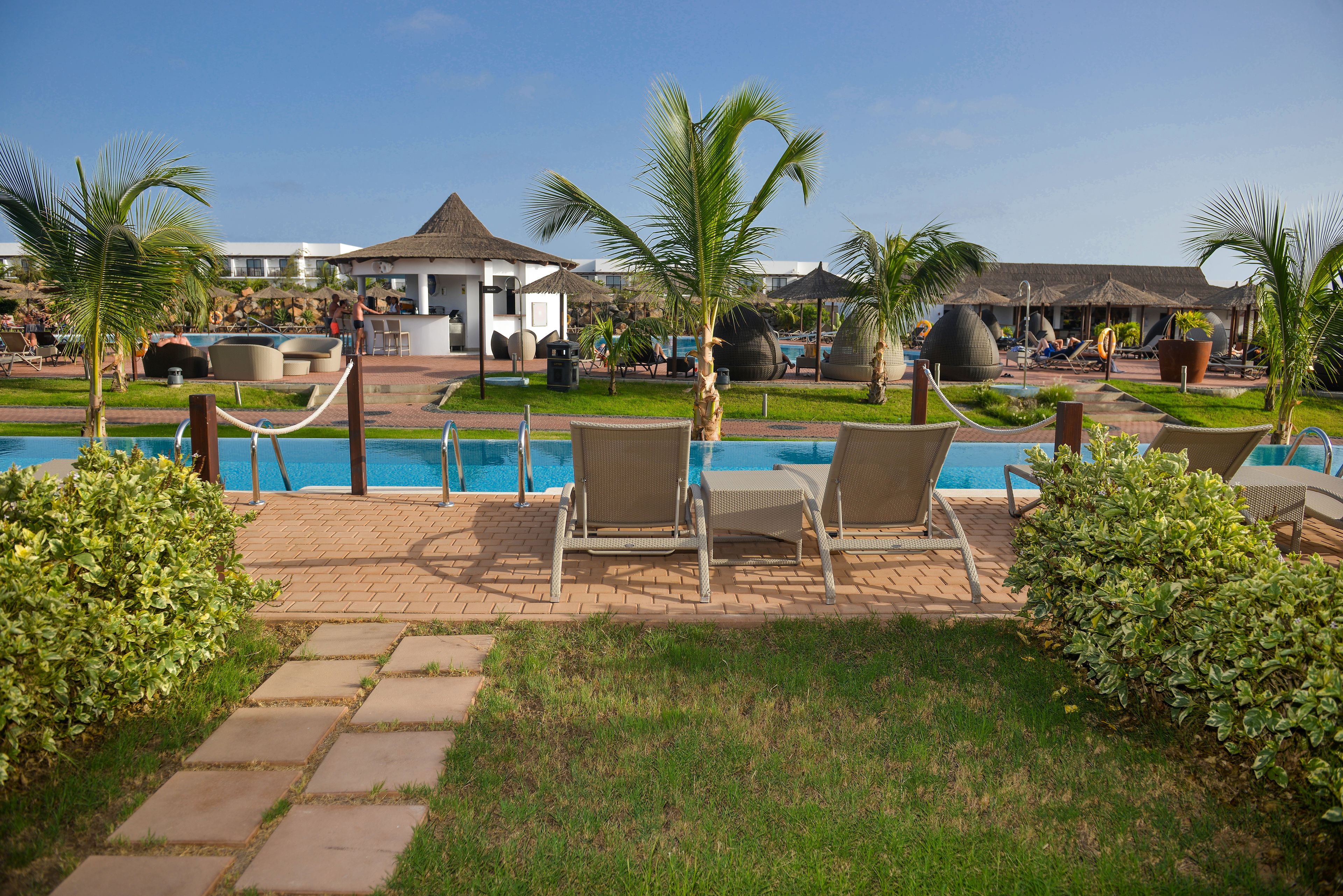 Un espace aménagé au bord de la piscine, avec des chaises longues, des palmiers et un bâtiment doté d’un toit de chaume.