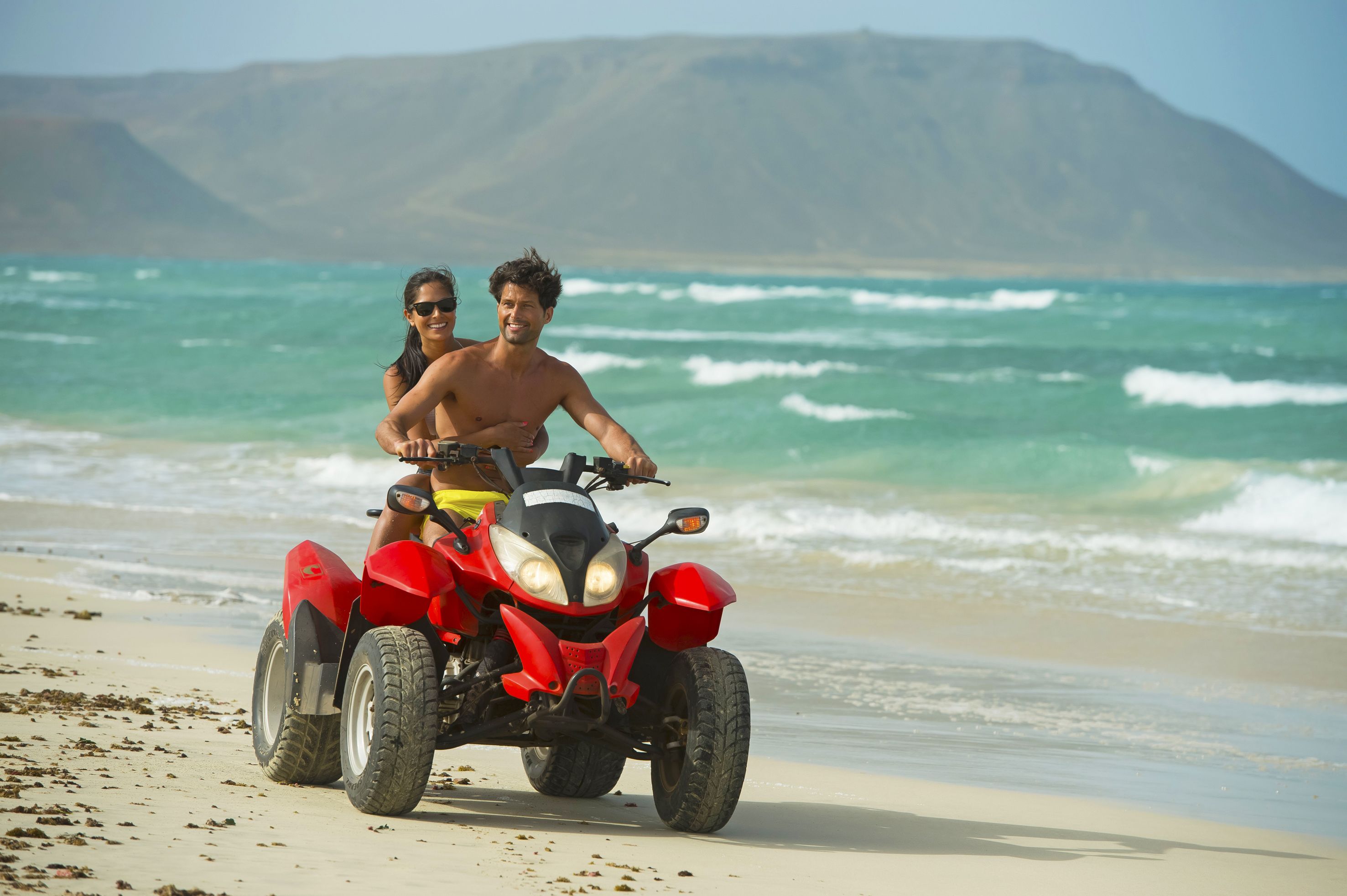 Un cuatrimoto rojo en una playa arenosa con olas del océano al fondo.