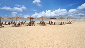 Ein Strand mit Liegestühlen, reetgedeckten Sonnenschirmen, einem Volleyballnetz und blauem Himmel.