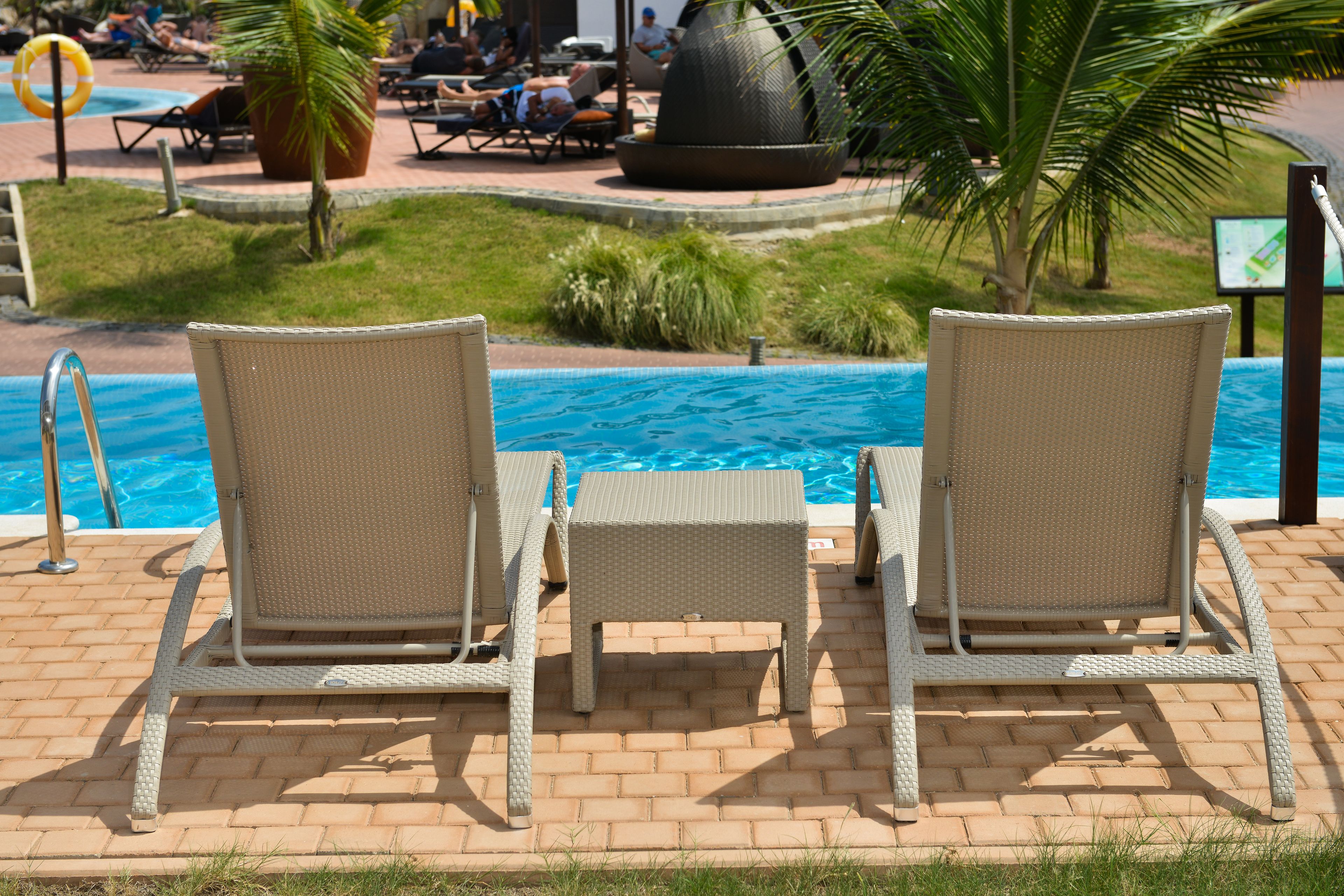A poolside area with two wicker lounge chairs and a small table, surrounded by a brick patio and palm trees.