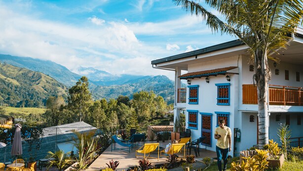 Un edificio de dos pisos con paredes exteriores azules y blancas, un balcón con barandillas naranjas y vistas a montañas al fondo.