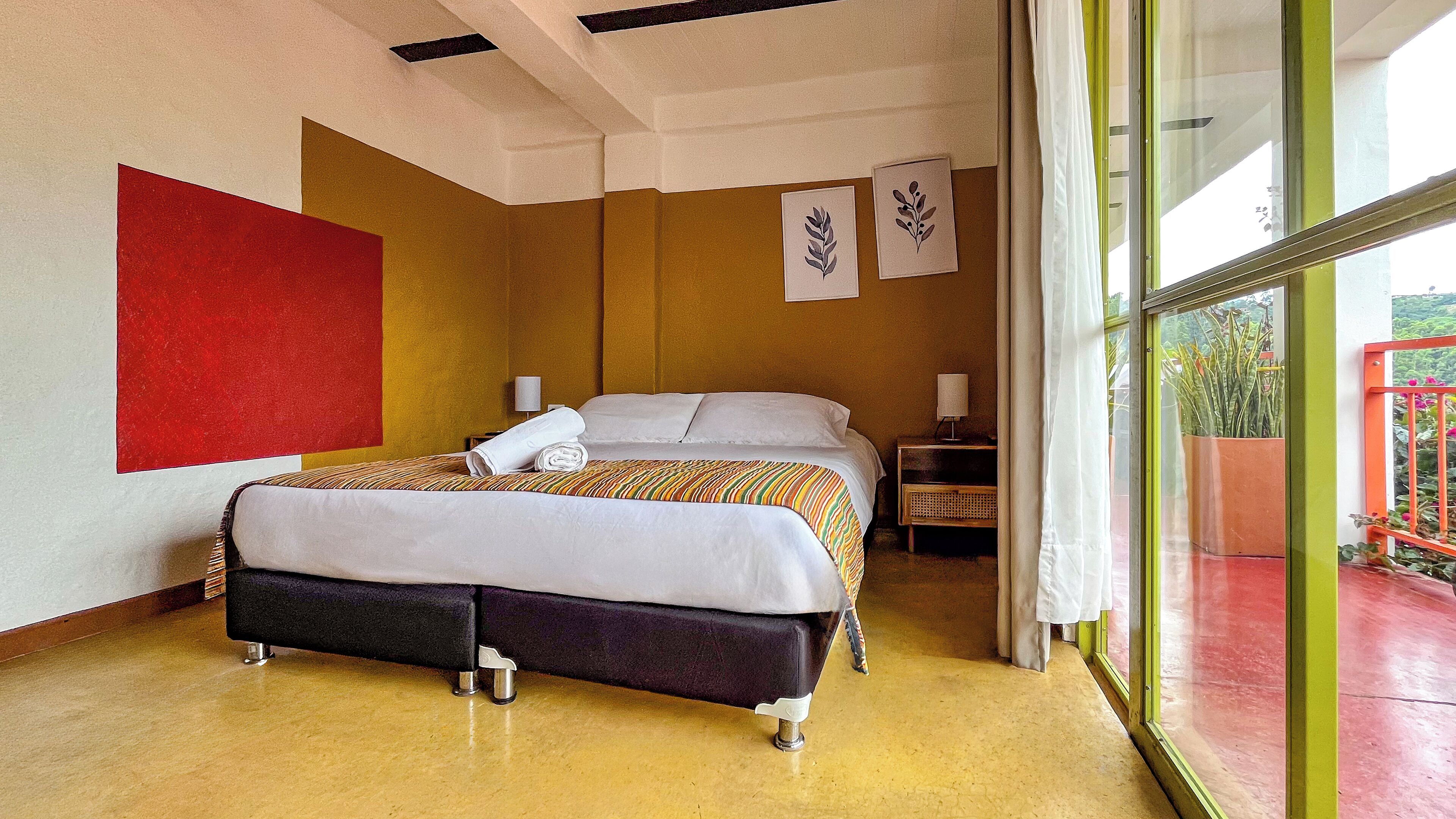 Una camera d'albergo moderna con un letto grande, un comodino, una lampada a parete e vista all'esterno attraverso un'ampia finestra.