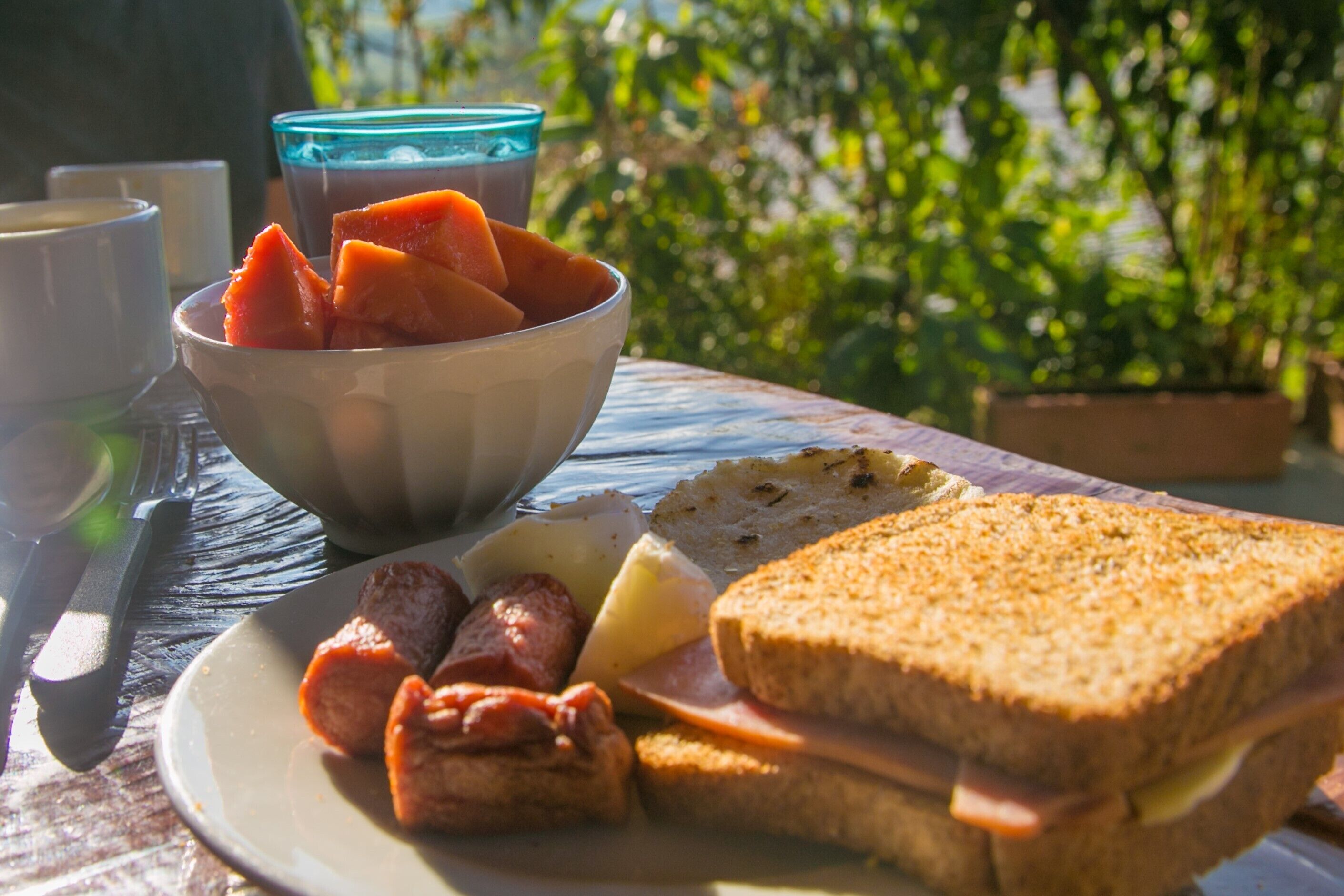 A plate with a sandwich, sausage, cheese, and a bowl of fruit.